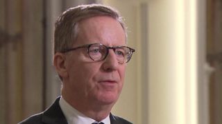 Ein älterer Mann mit kurzen, hellbraunen Haaren, der einen schwarzen Anzug, ein weißes Hemd und eine schwarze Krawatte trägt. Er trägt eine schwarze Brille mit klaren Gläsern. Er schaut direkt in die Kamera.