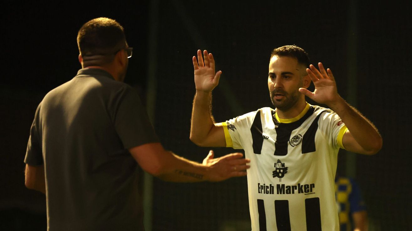 Ein Mann mit einer Tätowierung am Arm gestikuliert mit den Händen. Er trägt ein gestreiftes Trikot mit dem Namen und Logo von Erich Marker. Ein Mann mit Brille steht hinter ihm.