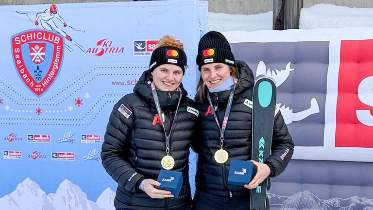 Two women pose for a photo, smiling, while holding medals and a trophy. They wear winter jackets and hats. The backdrop features a white banner with logos and text.