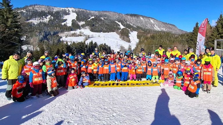 A large group of people in safety gear, including children, pose for a photo on a snowy mountain. A banner with the word Raiffeisen is in the foreground.