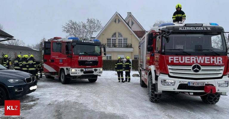 Zwei Feuerwehrleute stehen vor einem Haus mit geparkten Feuerwehrwagen im Schnee. Ein Feuerwehrmann ist auf einem der Trucks.