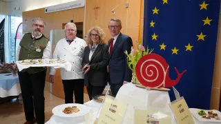 Vier Personen stehen an einem Tisch mit Speisen und Tellern. Der Mann links hält einen Teller, und die Frau lächelt. Der Mann in der Mitte trägt einen Anzug. Dahinter ist eine blaue Flagge mit Sternen.