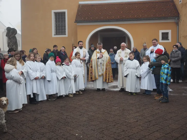 Priester und Kinder in weißen Gewändern versammeln sich vor einer Kirche. Die Priester halten Kerzen, und einige Kinder tragen bunte Mützen.