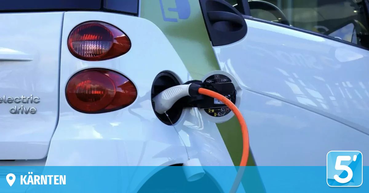 Ein weißer Elektrowagen wird mit einem orangefarbenen Kabel an einer Ladestation aufgeladen. Die Rücklichter des Autos sind rot.