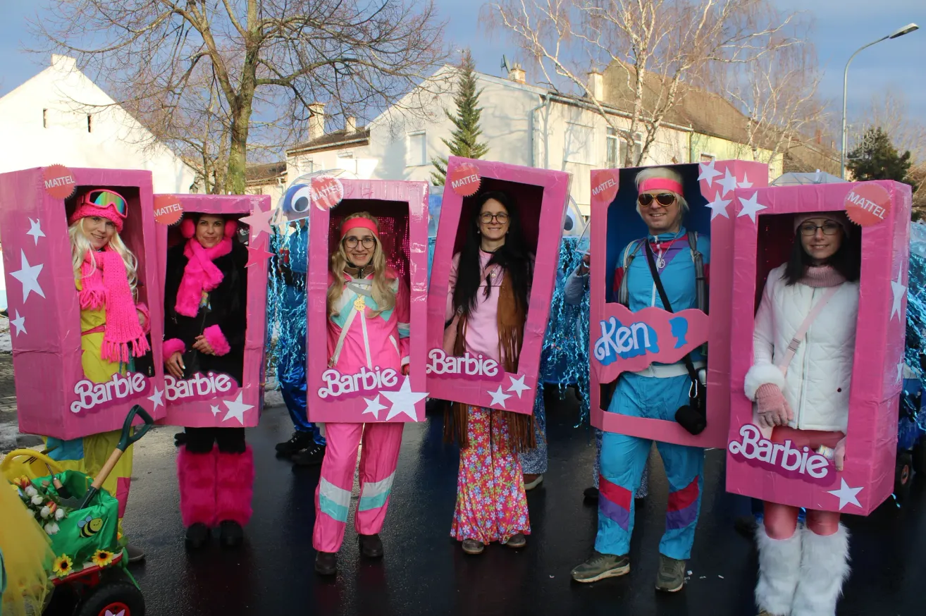 Eine Gruppe von Menschen, die in rosa Kostümen mit Barbie-Kisten auf dem Kopf und am Körper gekleidet sind. Sie stehen auf einer Straße, wahrscheinlich für eine Parade.
