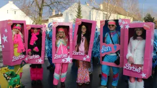 Eine Gruppe von Menschen, die in rosa Kostümen mit Barbie-Kisten auf dem Kopf und am Körper gekleidet sind. Sie stehen auf einer Straße, wahrscheinlich für eine Parade.