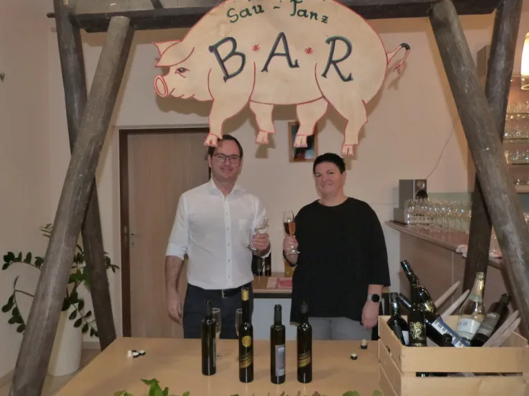 Two individuals stand behind a table holding wine glasses, with a pig sign reading 'Sau-Fanz BAR' above them. They are surrounded by wine bottles.
