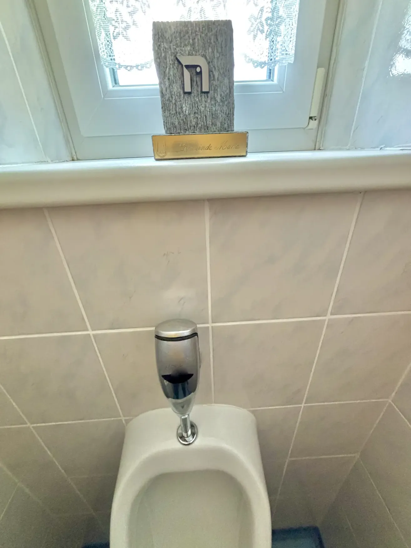 Ein weißer Urinal mit einem silbernen Spüler steht an einer gefliesten Wand. Ein goldenes Schild mit schwarzem Rand befindet sich darüber. Ein Fenster ist über dem Schild.