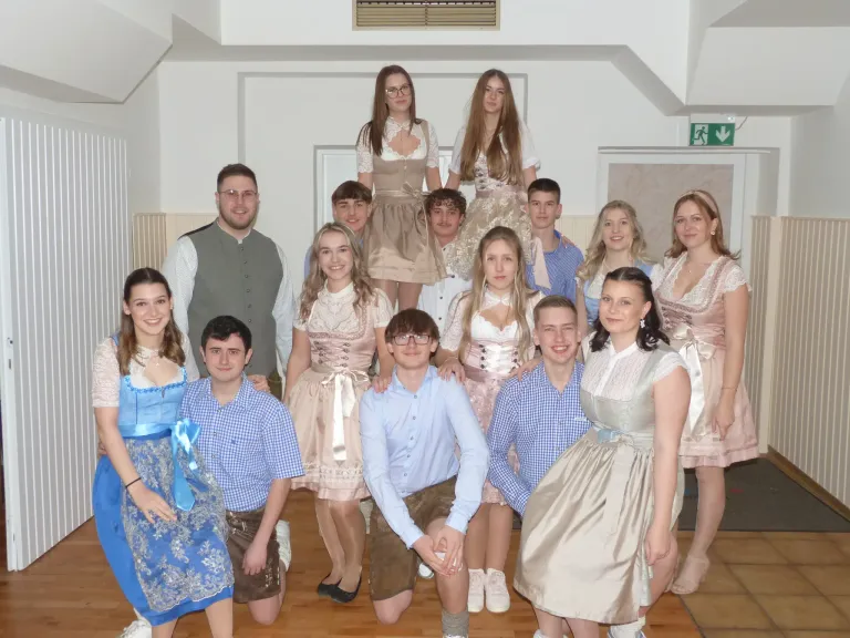 Eine Gruppe junger Menschen in traditioneller Kleidung posiert für ein Foto in einem Raum mit weißen Wänden und Holzboden. Die Frauen tragen Kleider und die Männer blaue Hemden. Zwei Frauen stehen auf den Schultern zweier Männer in der Mitte.
