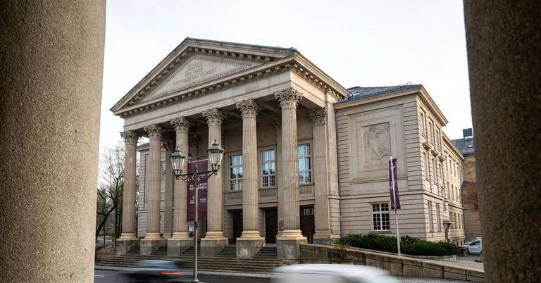 A classical architectural building with columns and a triangular pediment stands on a city street. Two street lamps are on either side of the entrance.