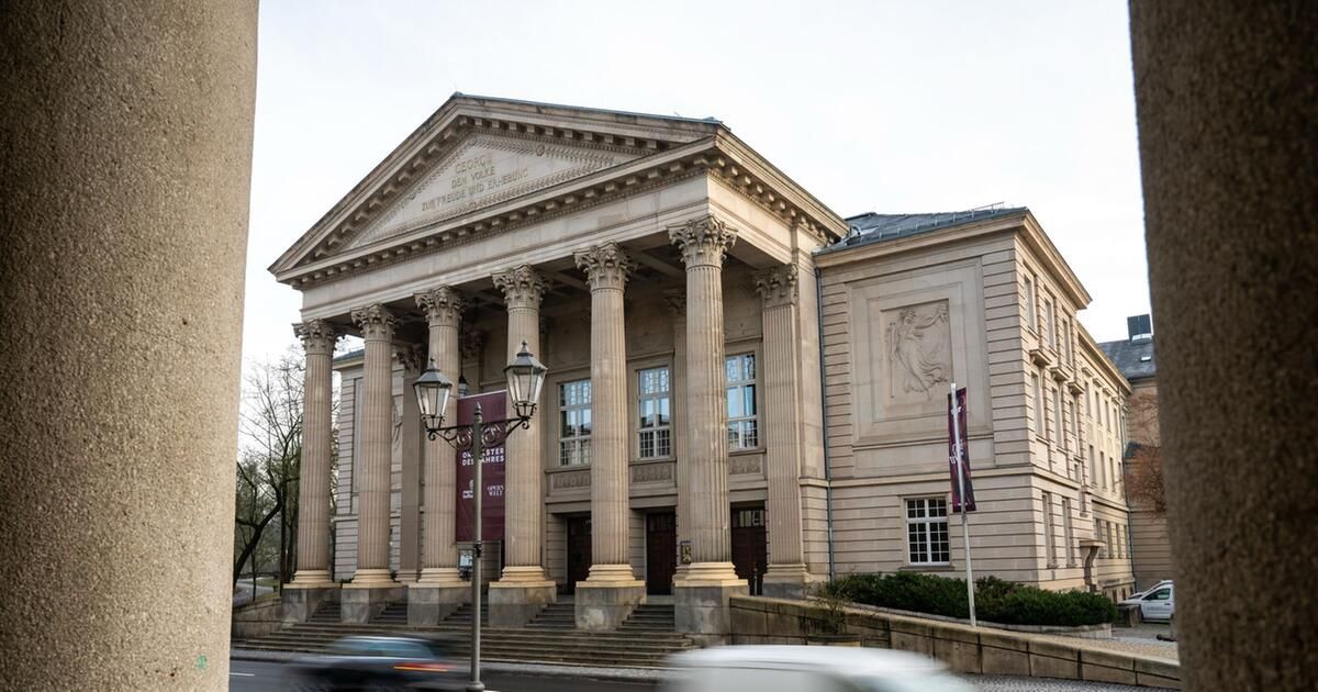 A classical architectural building with columns and a triangular pediment stands on a city street. Two street lamps are on either side of the entrance.