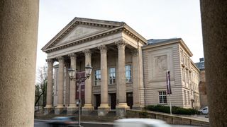 A classical architectural building with columns and a triangular pediment stands on a city street. Two street lamps are on either side of the entrance.