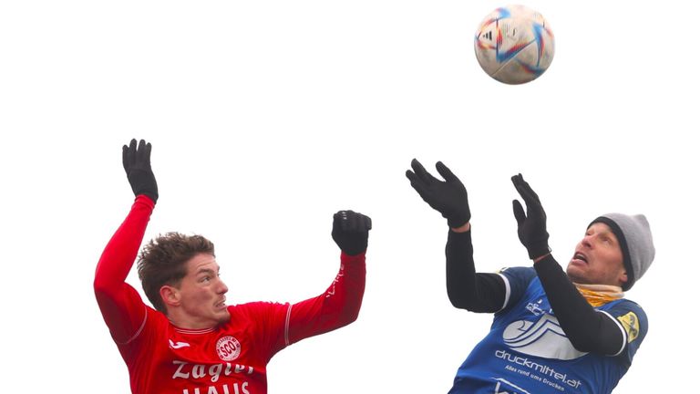 Two soccer players reach up for a ball in the air. One wears a red jersey with 'Zagwen' and the other a blue jersey with 'druckmi'.