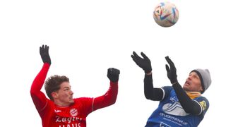 Two soccer players reach up for a ball in the air. One wears a red jersey with 'Zagwen' and the other a blue jersey with 'druckmi'.