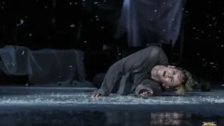 A person in a dark outfit lies on the floor, looking up in distress. Snowflakes float around them, and the background is dimly lit.