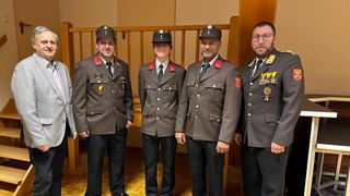 Four men in military uniforms, adorned with medals, stand in a row, smiling. They are positioned in front of a wooden railing, with a wooden wall and staircase behind them.