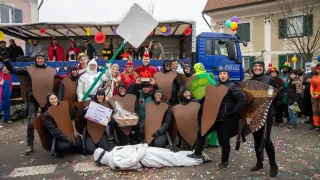 A group of people in costumes poses for a photo in front of a blue truck, a building, and confetti. Some wear masks, and one holds a sign.