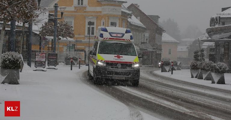 Ein Krankenwagen fährt mit eingeschalteten Lichtern durch eine verschneite Straße vor einem schneebedeckten Gebäude.