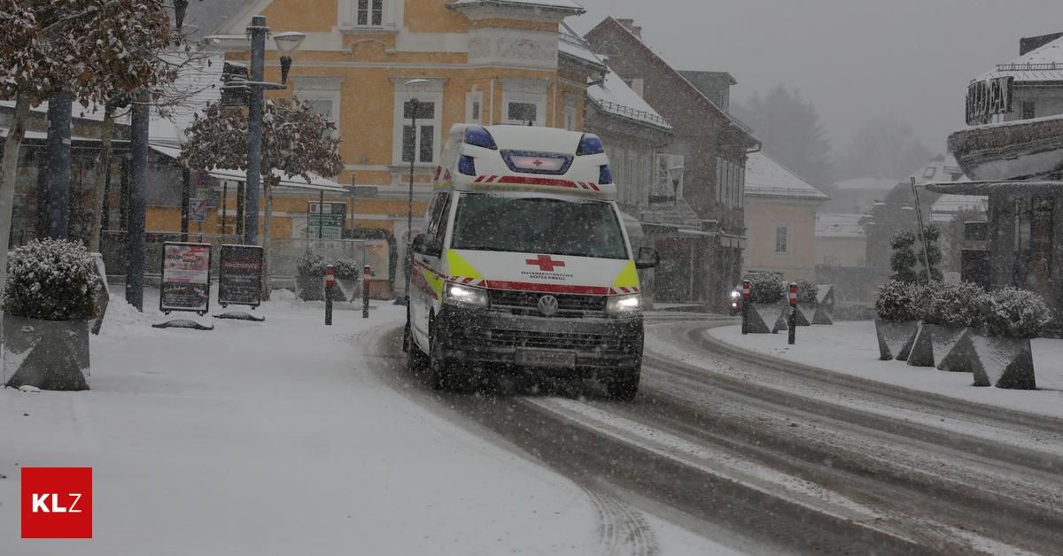 Ein Krankenwagen fährt mit eingeschalteten Lichtern durch eine verschneite Straße vor einem schneebedeckten Gebäude.