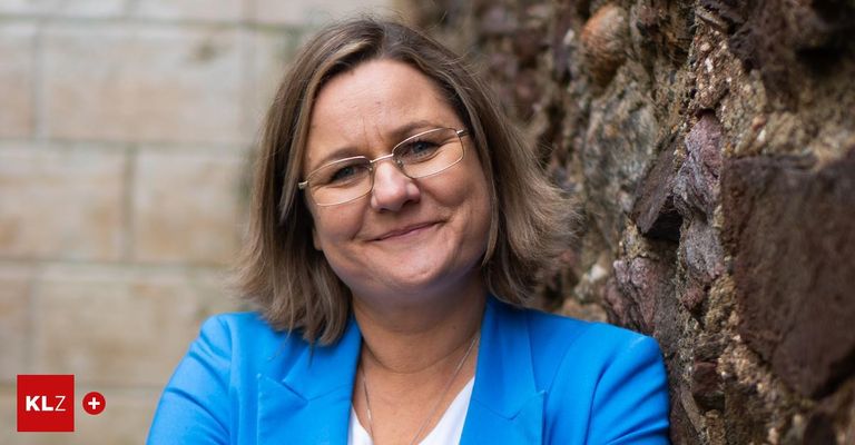 Eine Frau mit kurzen braunen Haaren und einer Brille, die einen blauen Blazer und eine Halskette trägt, lehnt sich gegen eine Steinmauer.