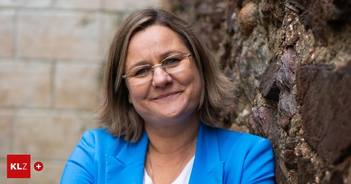 Eine Frau mit kurzen braunen Haaren und einer Brille, die einen blauen Blazer und eine Halskette trägt, lehnt sich gegen eine Steinmauer.