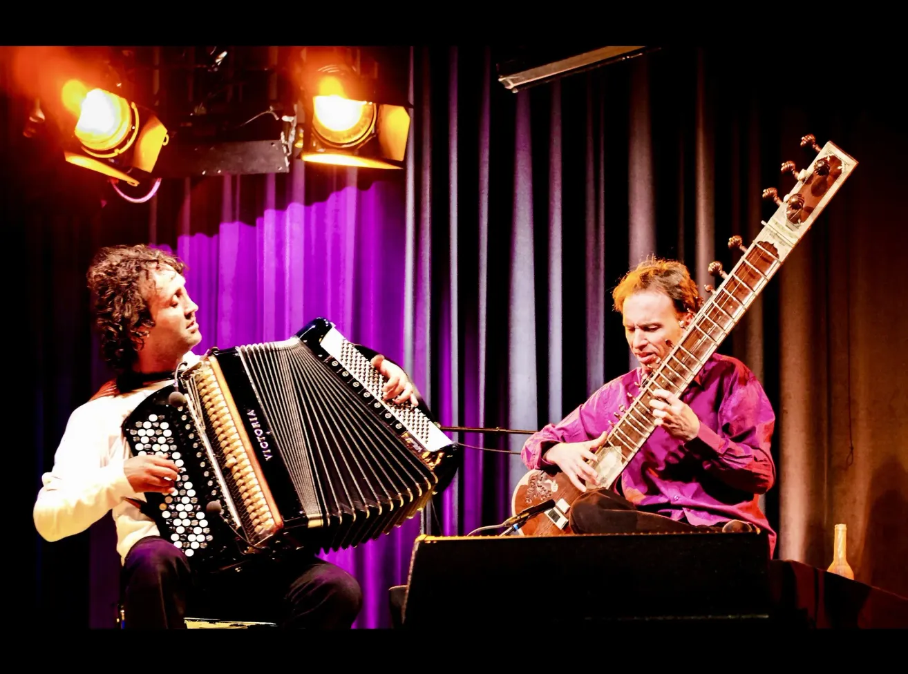 Two musicians are performing on stage. One is playing the sitar, and the other is playing the accordion. Spotlights shine on them.