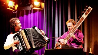 Two musicians are performing on stage. One is playing the sitar, and the other is playing the accordion. Spotlights shine on them.