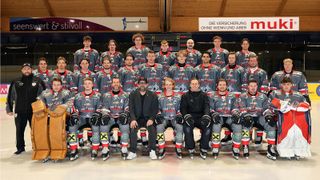 A hockey team is posing for a team photo with two coaches in front, all wearing jerseys and helmets. The wall behind has a banner with a sponsor's logo.