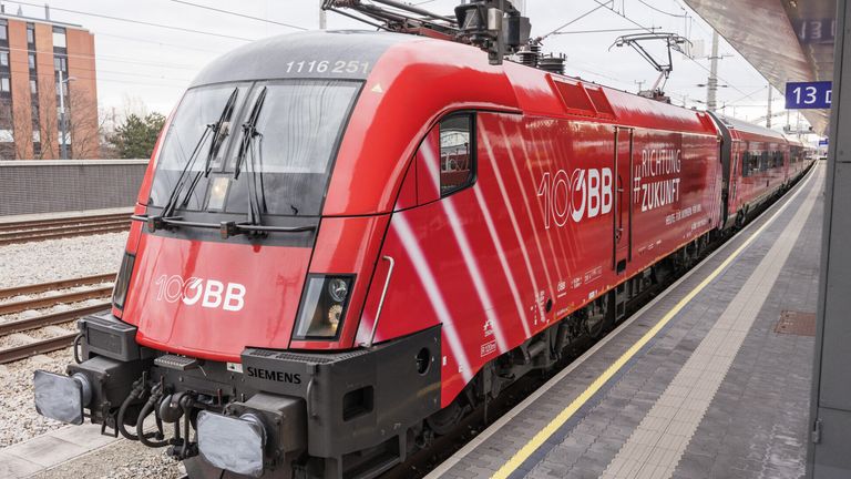 Ein roter Siemens-Zug mit der Nummer 1116251 steht auf einem Bahnsteig, wobei sich das verschwammte Bild im Frontfenster spiegelt. Er ist mit den Aufschriften '100BB' und 'OBB' gekennzeichnet.
