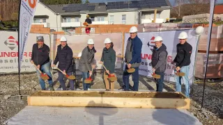 Group of people in construction gear are shoveling dirt into a wooden box in front of a building under construction.