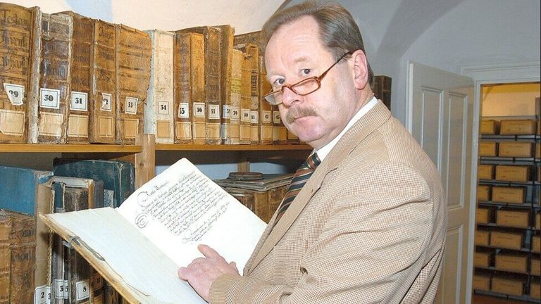 An older man in glasses and a suit is holding an old book in a library filled with numerous books on shelves.