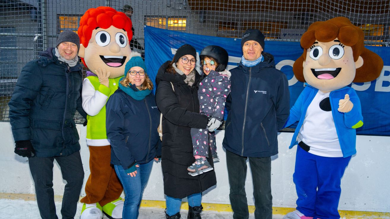 A family poses for a photo on an ice rink with two mascots, one wearing a red wig and the other green. The woman holds a child wearing a helmet, while the man wears a beanie.