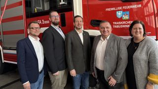 Four men stand next to a fire truck. They are all wearing suits. The truck has the words Freiwillige and Triasikbus on it. The man in the middle is wearing glasses.