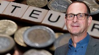A man stands in front of a pile of coins with a word board spelling 'TEU'.