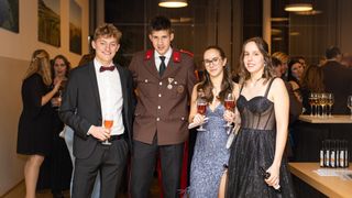 A group of young adults dressed in formal attire. A man in a military uniform stands between two women. They all hold wine glasses, and the man has a medal. The woman on the right holds a bottle of wine.