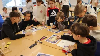 Several children are seated at a table with educational materials, engaged in an activity. A boy is looking down at a map while holding a pen. The table has papers, markers, and a cup.