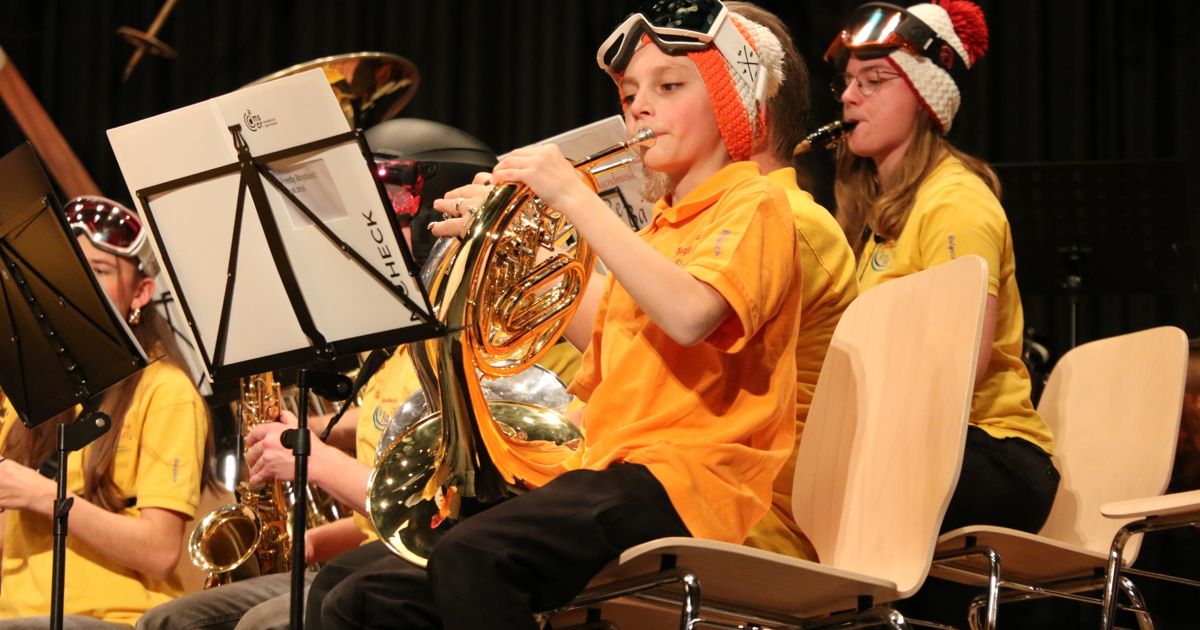 A child in an orange shirt plays a French horn while wearing goggles and a hat. Two other children sit nearby, playing saxophones, with a music stand in the background.