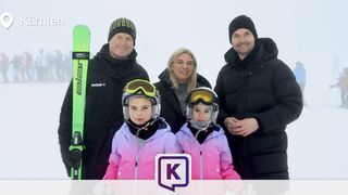 A family of four, including two young girls in ski gear, stands in the snow with smiles. The man on the left holds a green ski and the man on the right wears a black beanie.