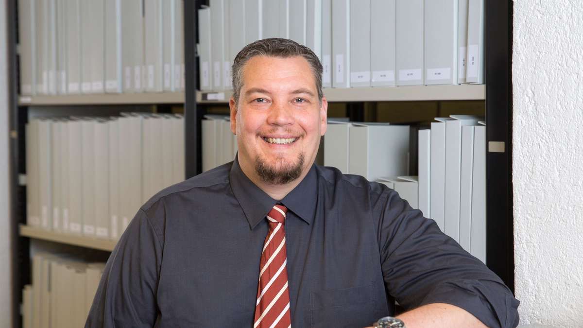 A man with a beard smiles in front of shelves filled with neatly organized white binders. He wears a dark blue shirt and a red and white striped tie.
