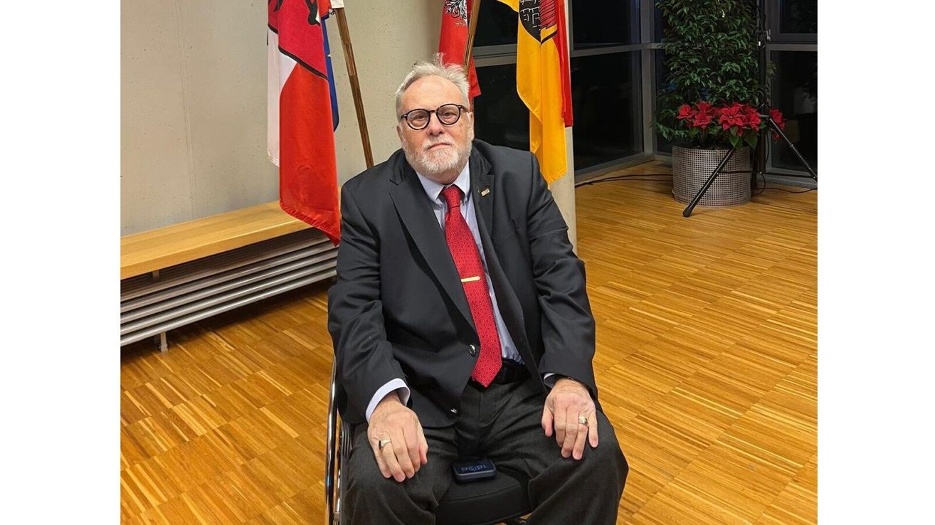 Ein Mann mit Brille und roter Krawatte sitzt in einem Rollstuhl vor Fahnen.