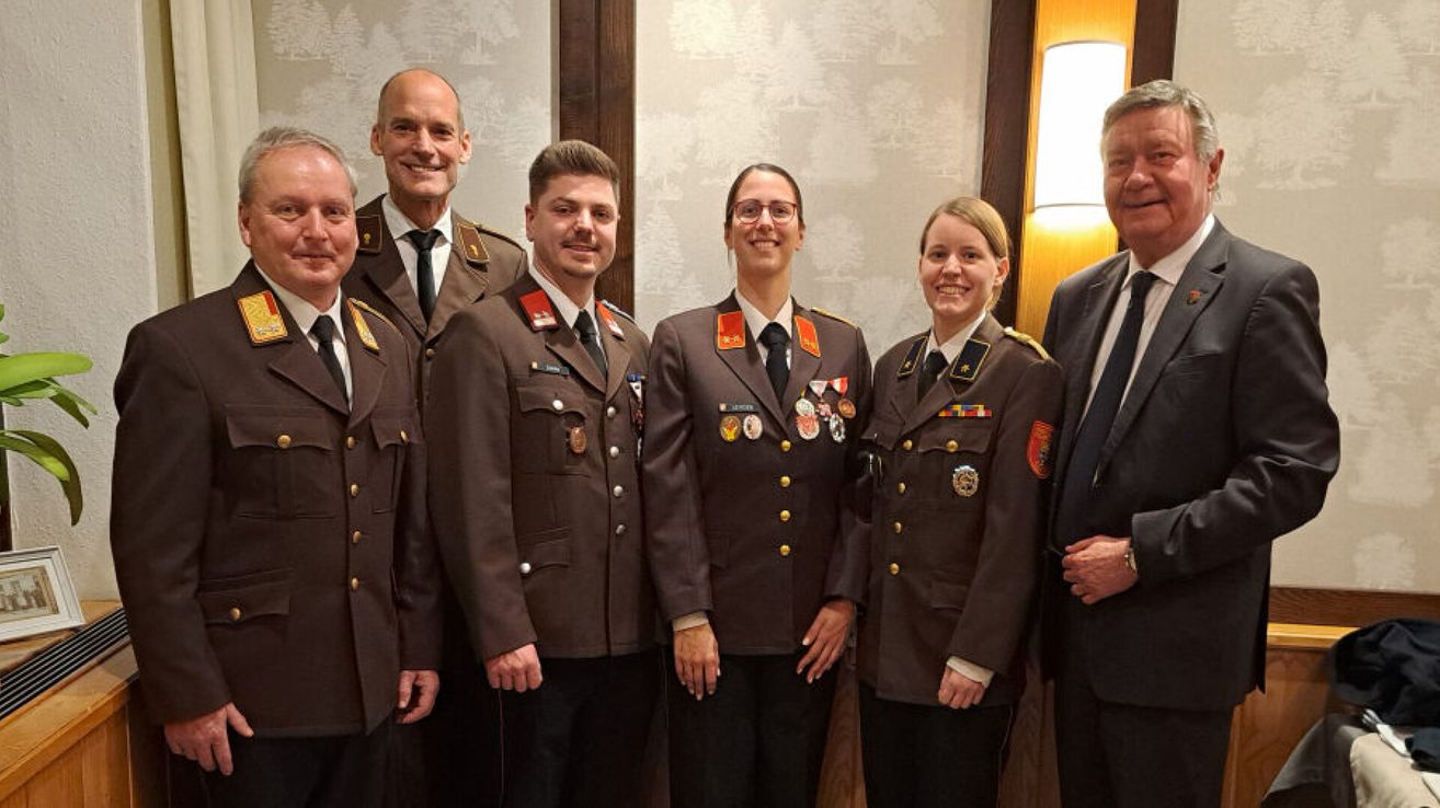 A group of people in military uniforms, including two women, stand together smiling. The background is a light-colored wall with a lamp.