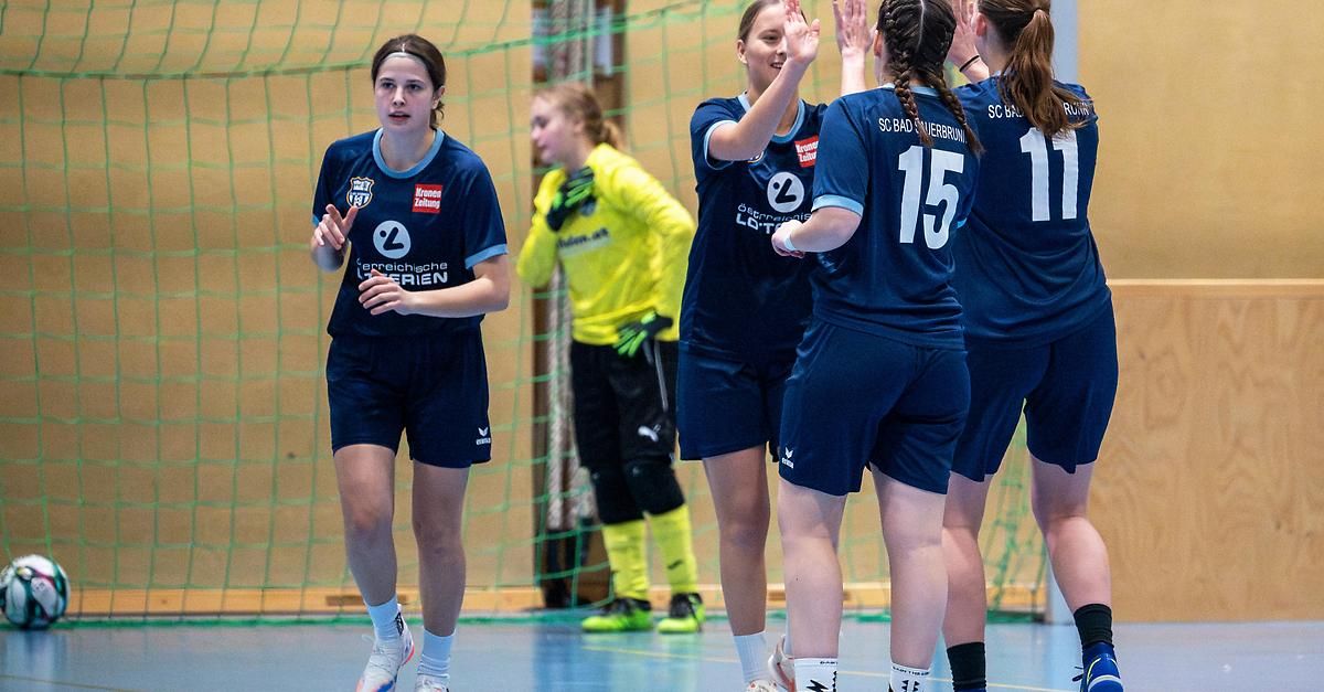 In an indoor sports arena, several women wearing sports uniforms are standing together. The woman in front is wearing a blue jersey with a logo. Another woman behind her is wearing a yellow jersey. They are probably celebrating a victory.