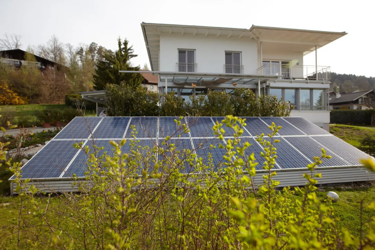 Ein modernes Haus mit Solarpanels vor einem klaren Himmel, umgeben von Grün und Bäumen.