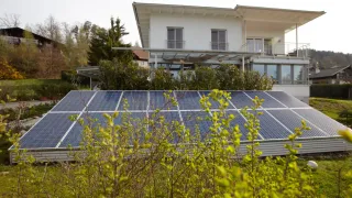 A modern house with solar panels in front, surrounded by greenery and trees under a clear sky.