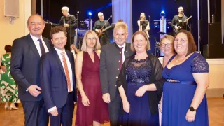 A group of adults, both men and women, are posing for a photo on a stage. They are all smiling. Behind them, musicians are playing instruments.