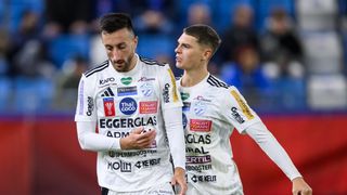 Two soccer players in white uniforms are on a field. One has 'EGGERGLAS' on his shirt, while the other has 'FAULST ANN'. They appear to be conversing. Behind them, a blurred crowd is watching.