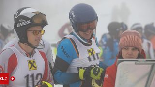 A skier with a blue helmet and goggles smiles at the camera, holding a ski pole. Nearby, two other people in ski gear watch, one wearing a white and red jacket with '112' on it.