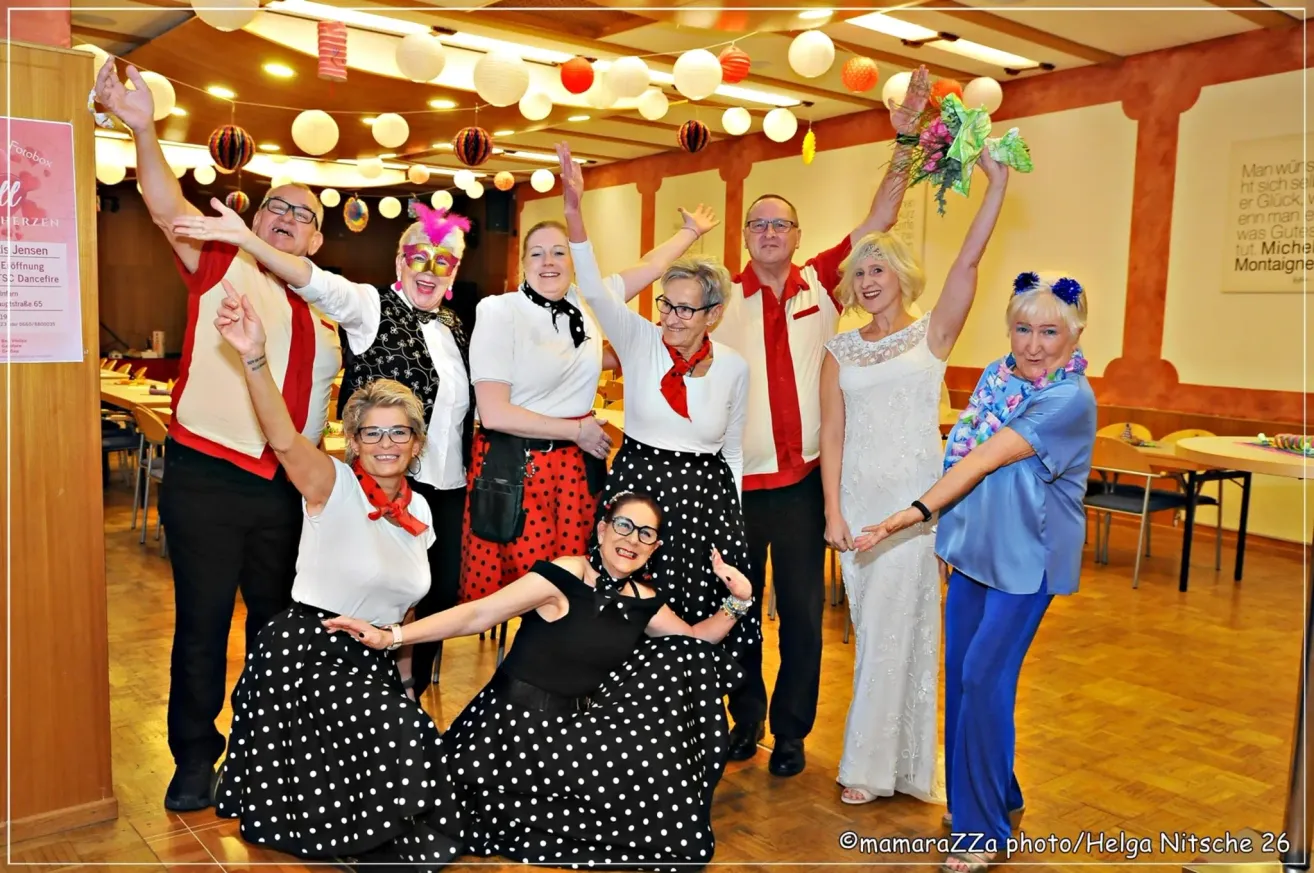 A group of adults dressed in themed attire, including polka dots and red tops, are celebrating with raised hands and festive decorations in the background.