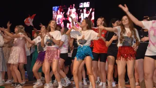A group of women dancing on a stage with a large screen in the background. They wear colorful skirts, white shirts, and sneakers. The screen shows a group of people.
