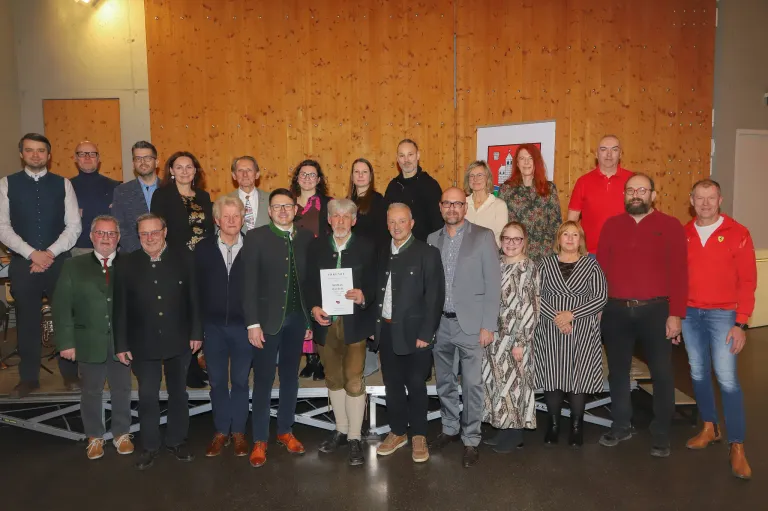 A group of people dressed in formal attire stand on a stage with a wooden wall. A man in the center holds a certificate. Behind them, a banner is mounted on the wall.